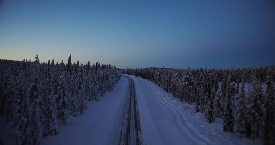 An aerial drone shot ascends from a snow-covered road to reveal a vast, remote forest landscape at dusk in winter