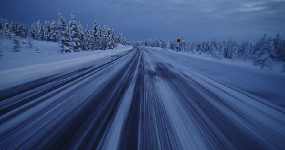An aerial drone flies quickly down a snow-covered road through a vast, wintry forest at dusk