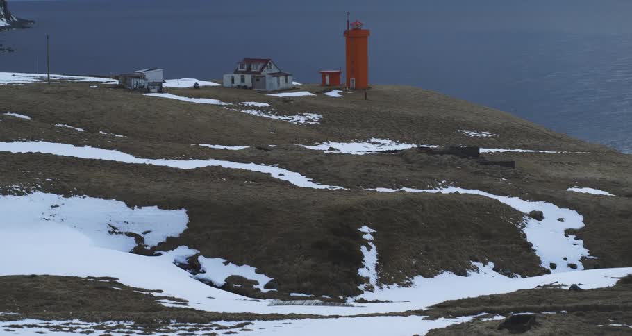 A flock of sheep walks through a desolate, snowy landscape with a bright orange lighthouse overlooking the ocean in Iceland