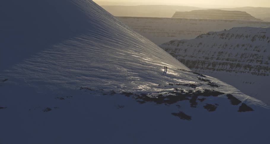 Static aerial shot of two people hiking along a vast, snowy mountain pass during a beautiful sunset