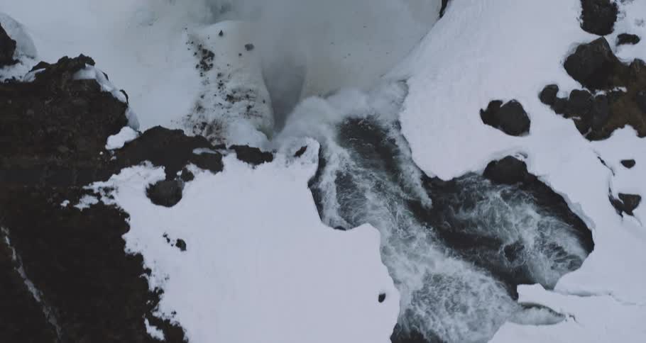 A dramatic top-down aerial shot follows a rushing river through a snow-covered landscape as it disappears into a steamy chasm