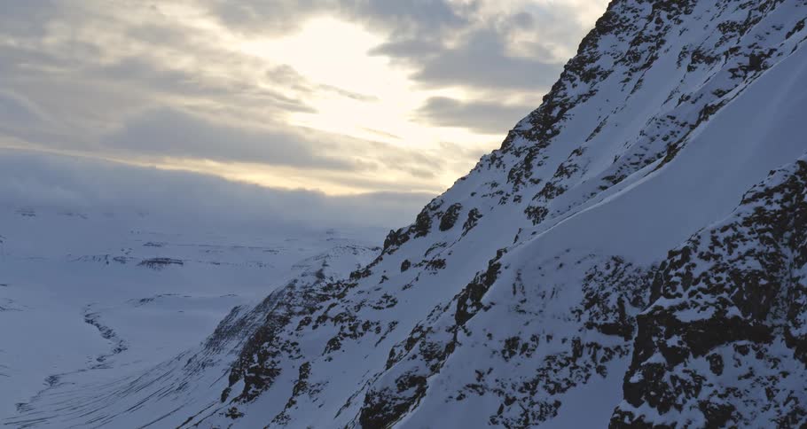 The sun glows behind thick clouds over a majestic, snow-covered mountain and a vast, frozen valley
