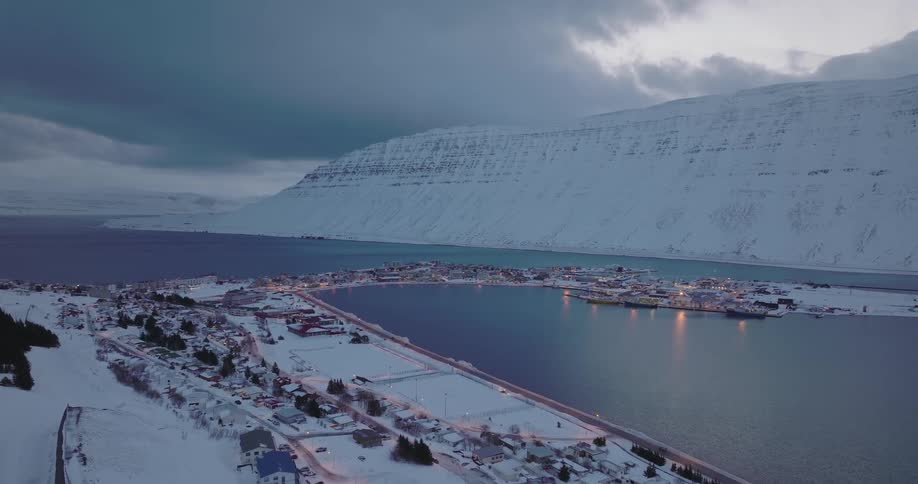 A tranquil aerial view shows a snow-covered fishing village nestled in a massive fjord during a moody winter twilight in Iceland
