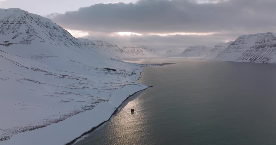 A stunning wide aerial view of a snowy fjord and mountains with sunlight breaking through the clouds