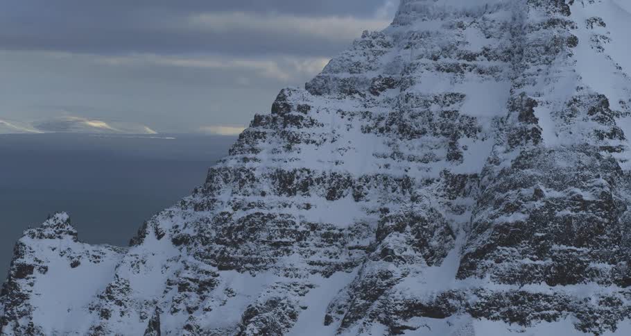 A majestic snow-covered mountain looms over a calm sea under a cloudy winter sky