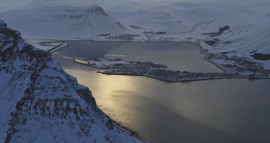 A serene, high-angle view of a coastal town in a snowy fjord as golden sunlight reflects on the calm water