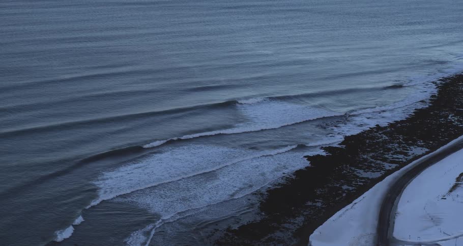 A wide, high-angle view of the cold ocean waves meeting the snow-covered shoreline of a remote coast