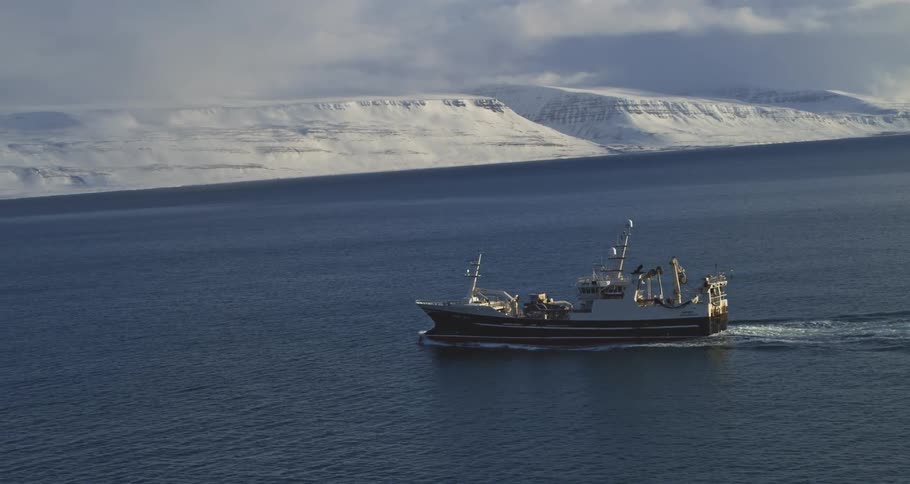 A large fishing trawler sails through the cold arctic sea with a vast snow-covered mountain range in the background
