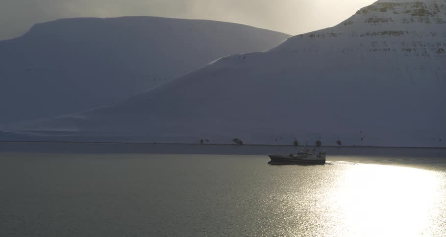 A fishing boat sails through a tranquil fjord at sunrise, surrounded by vast, snow-covered mountains