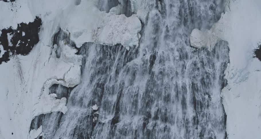 A top-down view captures a powerful waterfall cascading down dark rocks amidst a snowy landscape
