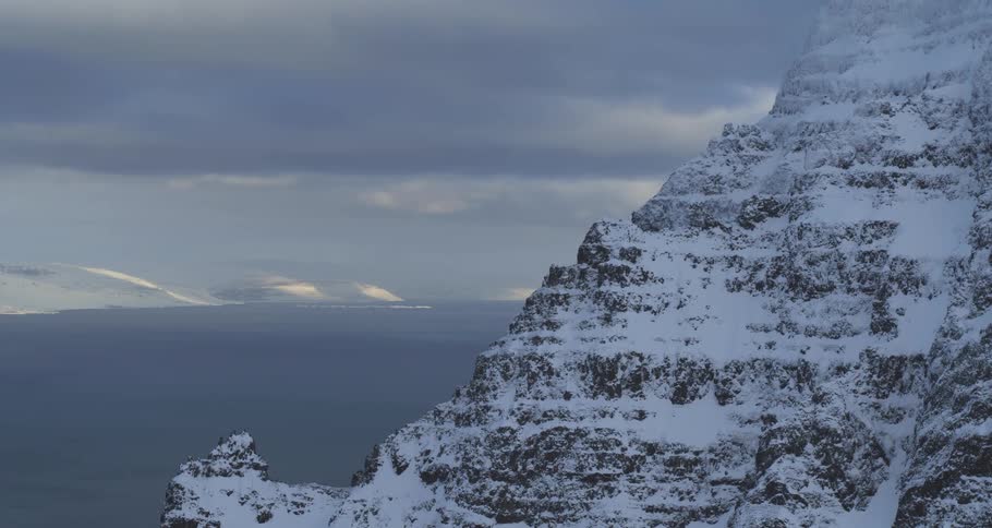 A vast winter landscape unfolds from a snowy mountain cliff, revealing a fjord and distant sunlit hills under an overcast sky