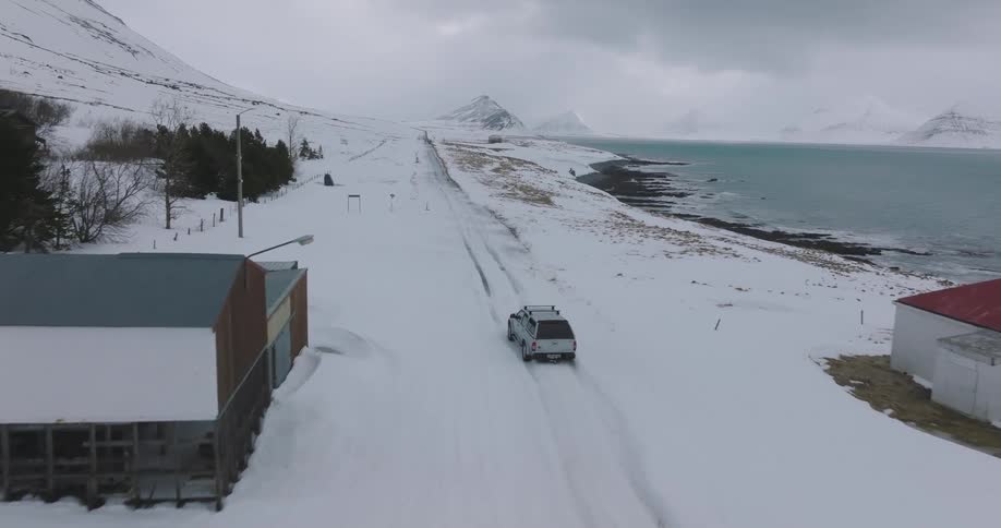 Aerial view of a silver pickup truck driving down a remote, snow-covered coastal road in a stark winter landscape