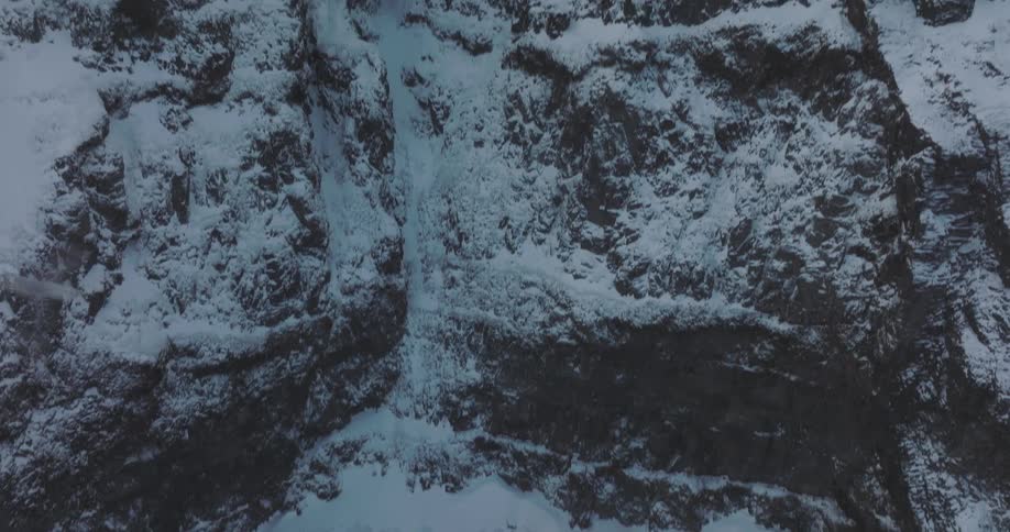 An aerial shot starts looking straight down at a snowy mountain crevice before tilting up to reveal the vast, rugged peak against an overcast sky