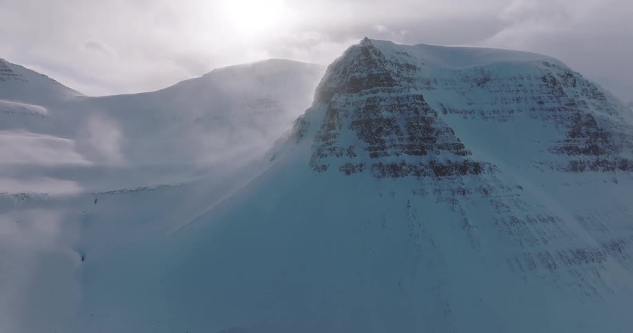 An epic aerial shot pulls back to reveal a vast, snow-covered mountain landscape with strong winds blowing snow across the slopes under a bright, cloudy sky