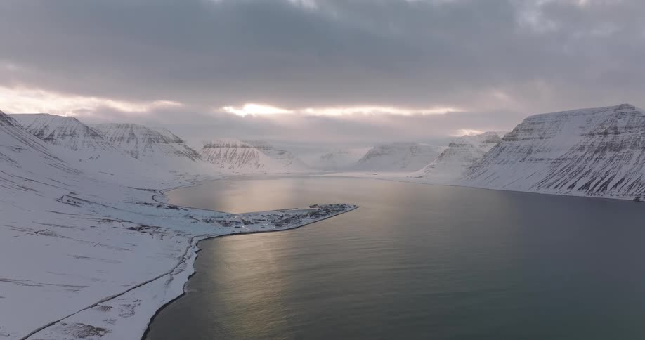 A still aerial shot reveals a vast, serene fjord with a small coastal town nestled between snow-covered mountains under a dramatic cloudy sky