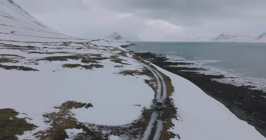 Drone follows a solitary van driving on a remote, snow-covered coastal road between mountains and the sea