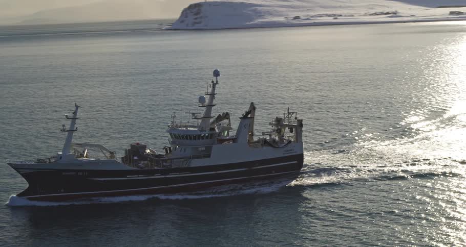 An aerial view captures a fishing trawler cruising through calm arctic waters past a snow-covered coastline at sunrise