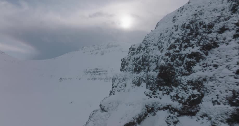 A cinematic aerial shot slowly reveals a stunning, snow-covered rock pinnacle in a vast arctic mountain range under an overcast sun