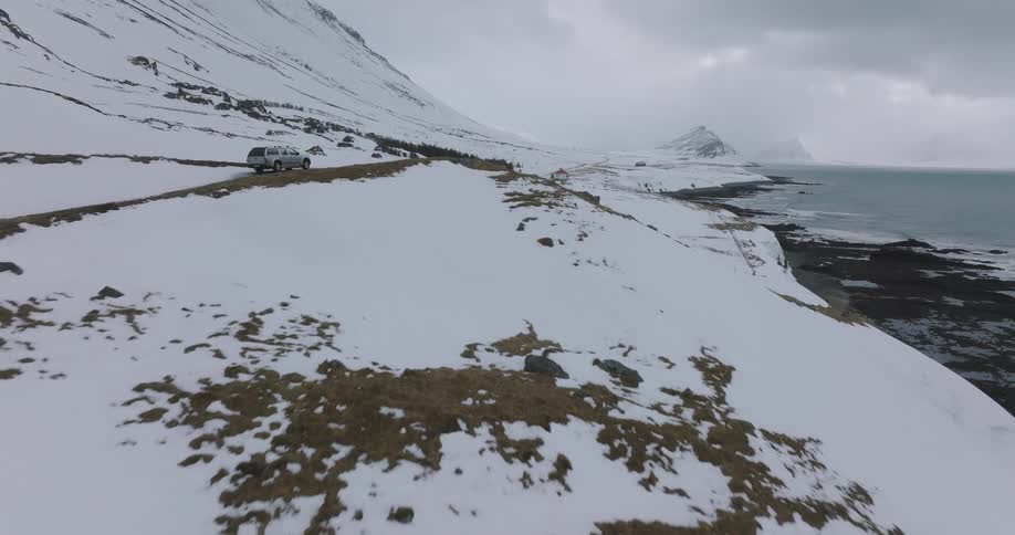 An aerial view follows a silver SUV driving along a remote coastal road through a vast, snow-covered mountainous landscape in winter