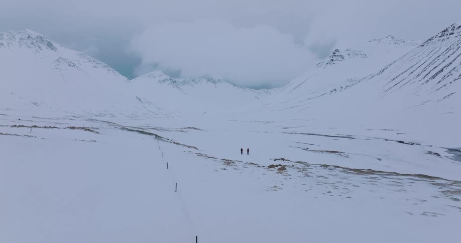 An epic aerial shot reveals two distant hikers walking through a vast, snow-covered mountain valley