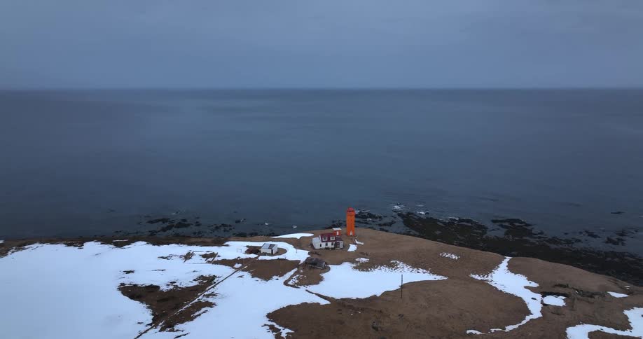Static aerial shot of a solitary orange lighthouse on a snow-dusted shoreline overlooking the calm, grey sea at dusk