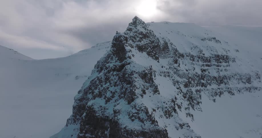 A slow aerial orbit reveals a formidable snow-covered mountain peak standing alone in a vast, desolate winter landscape