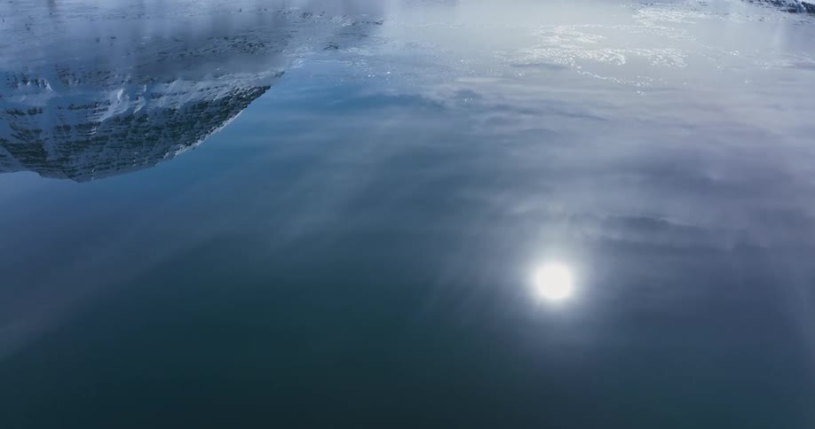 A stunning inverted reflection of a snowy mountain in a frozen lake flips to a fast aerial flight skimming across the icy surface