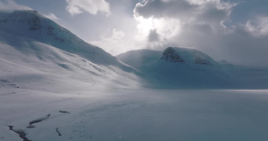 Dramatic aerial view of a desolate, snow-covered mountain valley with strong winds creating a ground blizzard under a moody, sunlit sky