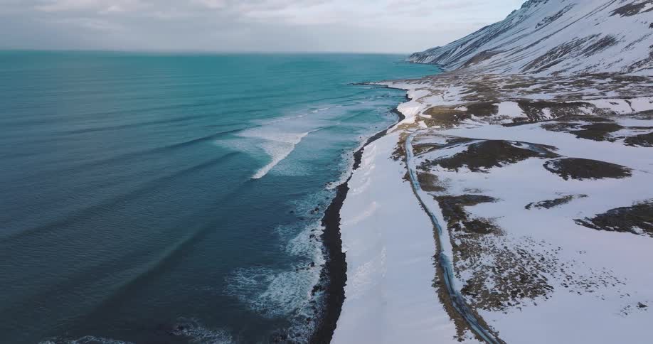 An aerial view captures the stunning contrast of turquoise ocean waves meeting a snow-covered coastline at the base of a mountain