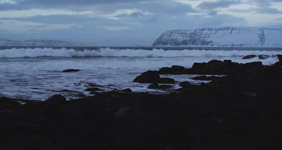A slow push-in shot reveals powerful ocean waves crashing on a dark, rocky shore in front of snow-capped mountains at dusk
