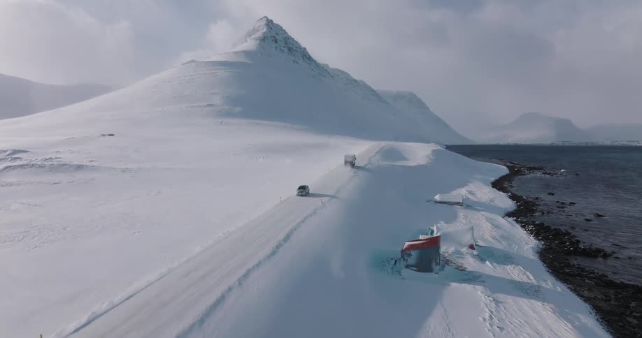 An aerial view shows cars driving on a remote, snow-covered coastal highway at the base of majestic mountains