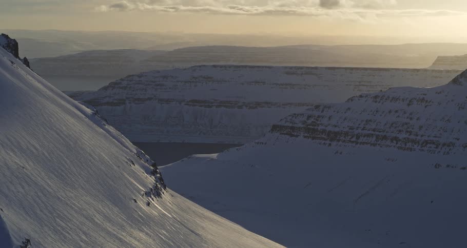 An aerial view slowly reveals a majestic, snow-covered mountain valley leading to a fjord at sunrise