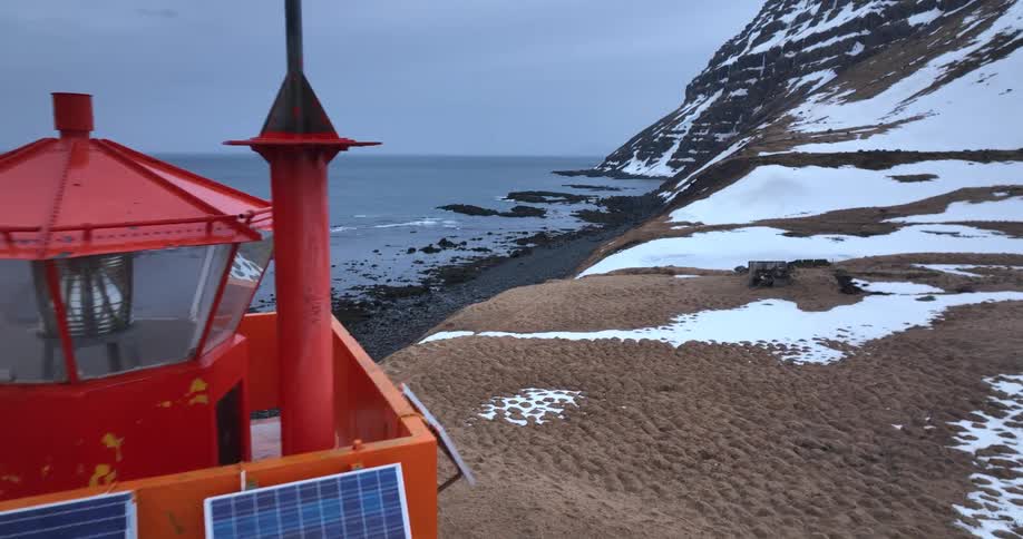 Drone footage captures a close-up of a remote orange lighthouse with solar panels on a snowy Icelandic coastline