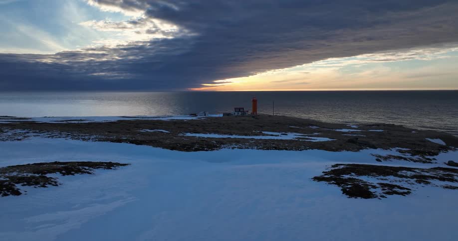 A dramatic aerial view shows an orange lighthouse on a snowy coast, with the sky split between dark storm clouds and a beautiful sunset over the ocean