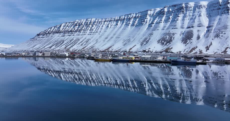 A picturesque Icelandic fishing village and massive snow-covered mountain range are perfectly reflected in the still, glassy fjord waters