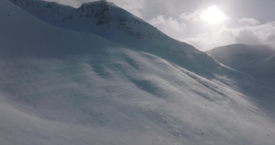 Blowing snow creates a ground blizzard across a desolate mountain range with the bright sun behind clouds