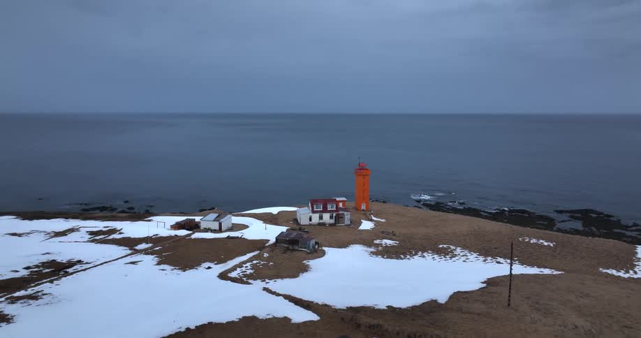 A striking orange lighthouse stands sentinel on a remote, snowy peninsula overlooking the vast, grey sea on an overcast day