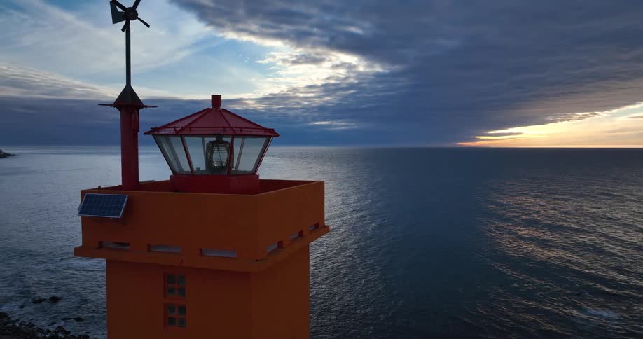 A dramatic aerial shot pulls back from an orange lighthouse overlooking the vast ocean at sunset on a snowy coastline