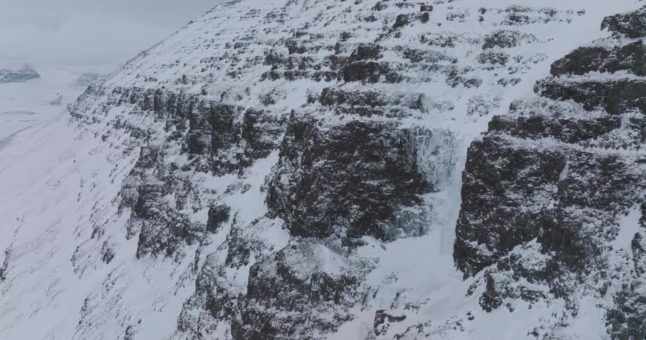 A slow aerial pullback reveals a massive frozen waterfall on a desolate snow-covered mountain