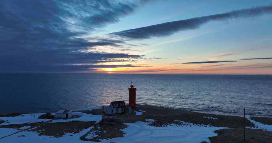 A slow aerial pullback reveals a striking red lighthouse on a snowy coast as the sun sets over the vast ocean