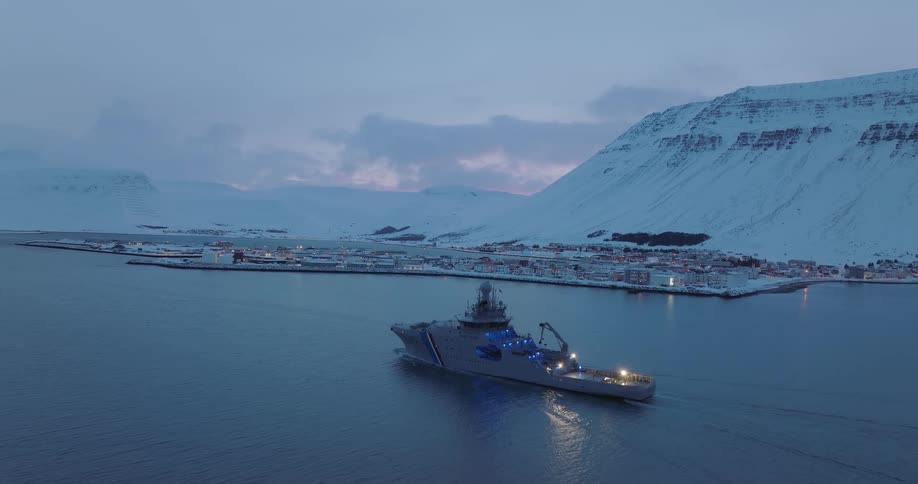 An Icelandic Coast Guard ship sails through a fjord at twilight with a snowy mountain village in the background