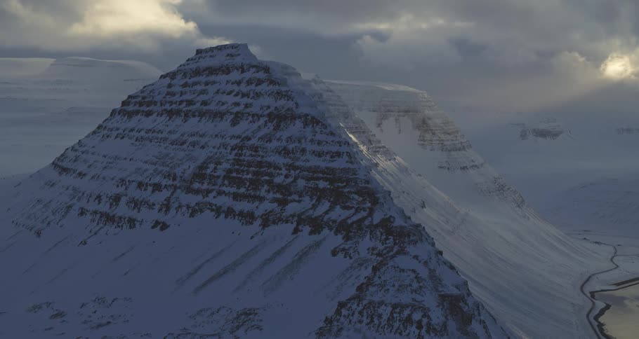 A majestic, snow-covered mountain stands tall under a dramatic cloudy sky with a sliver of sunlight breaking through