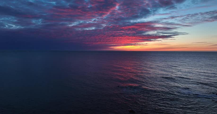 A breathtaking static aerial view of the ocean at sunrise, with vibrant pink and purple clouds reflecting on the calm water's surface