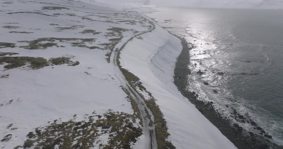 An aerial shot follows an SUV as it drives down a treacherous snowy road along a dramatic winter coastline