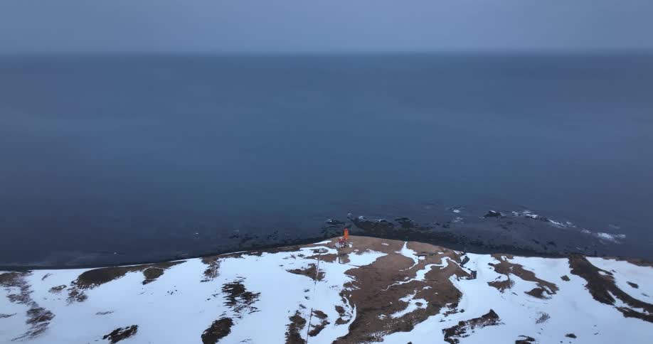 A static aerial view of a person on a snowy headland by a small orange lighthouse overlooking the vast ocean