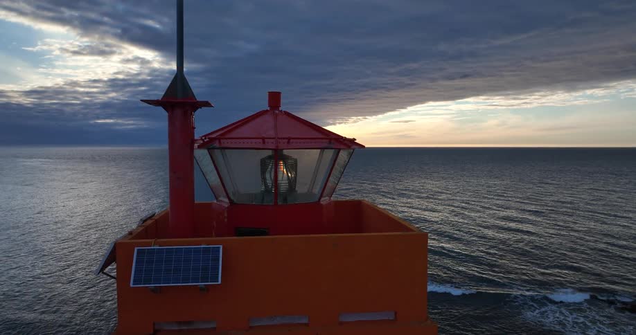 An orange lighthouse beacon rotates at sunset, overlooking a vast, dark ocean under a cloudy sky