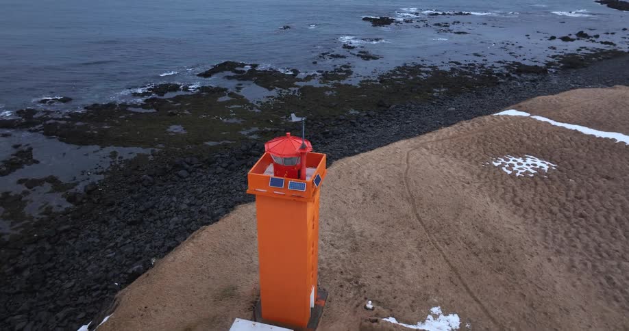 An aerial shot slowly approaches a vibrant orange lighthouse standing on a desolate, grassy cliff overlooking a dark, rocky sea