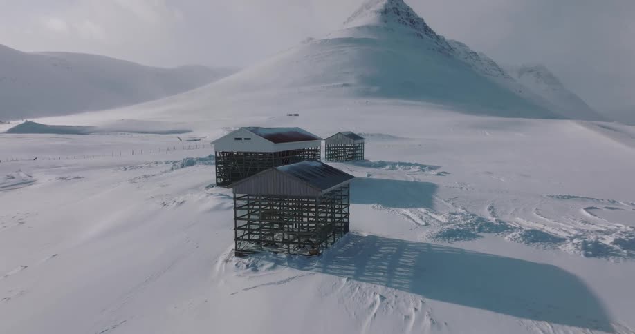 Aerial pullback shot reveals traditional fish drying racks in front of the iconic Kirkjufell mountain in a vast, snowy Icelandic landscape