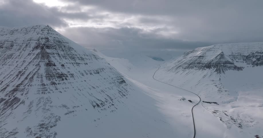A breathtaking aerial view captures a winding road through a vast, snow-covered mountain valley under a dramatic, cloudy sky