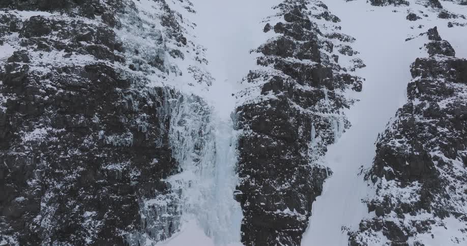A slow aerial ascent reveals a magnificent frozen waterfall nestled between rugged, snow-covered mountain peaks